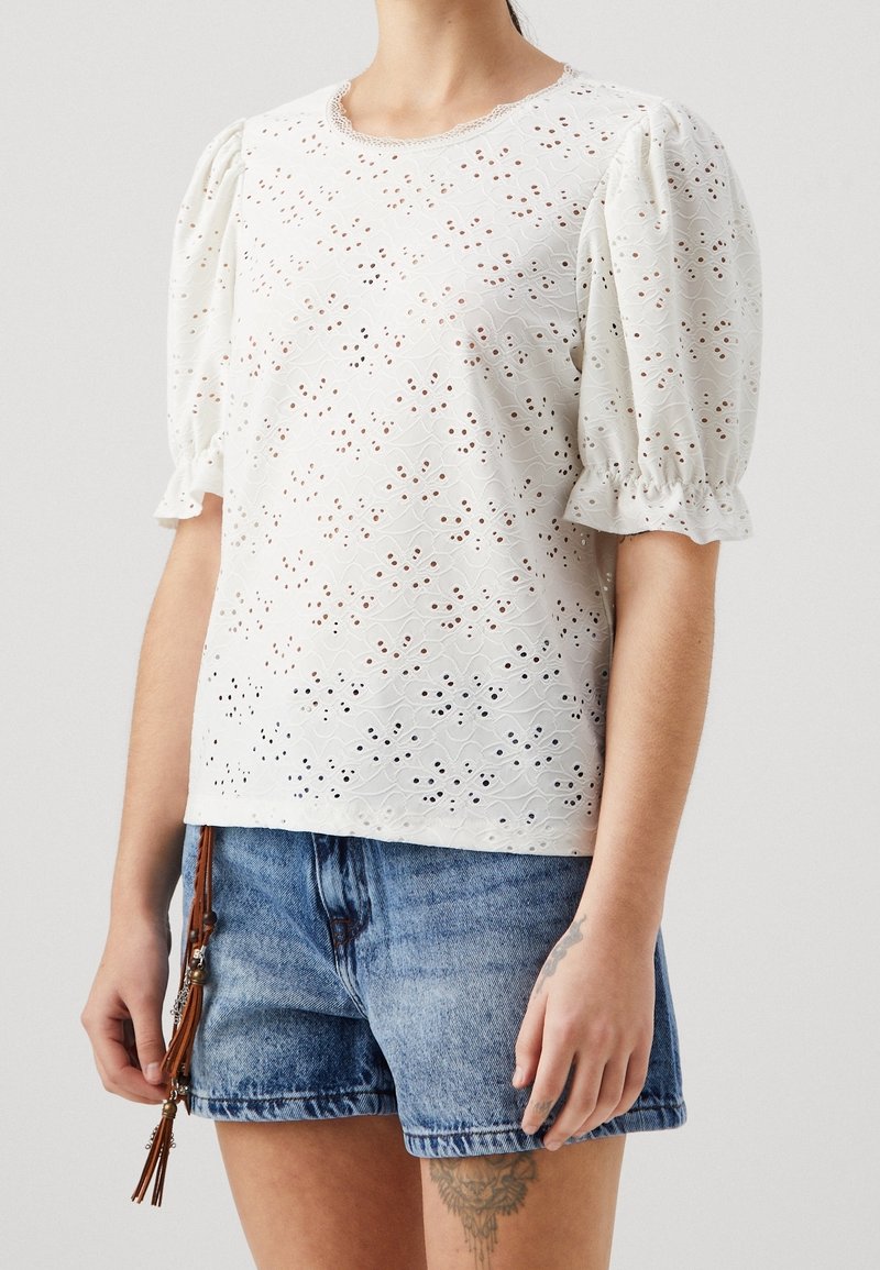 Femme portant un haut à manches bouffantes en broderie anglaise blanche et un short en jean bleu avec un accessoire à pampille marron suspendu à la taille.