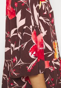 Détail de la manche en tissu marron avec imprimé floral rouge, rose et jaune et poignet élastique froncé sur un vêtement porté par une personne.