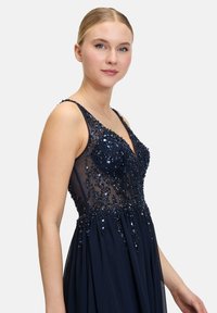 Granatowa wieczorowa suknia z cekinowym gorsetem i przezroczystymi detalami. Zwiewna spódnica, regulowane ramiączka oraz misternie wykonane ozdoby dla dodatkowej tekstury.