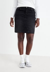 Černá denimová sukně s mírně třepeným lemem, předním zapínáním na patentky a diagonálními švy, doplněná šedými sportovními botami a bílými ponožkami.