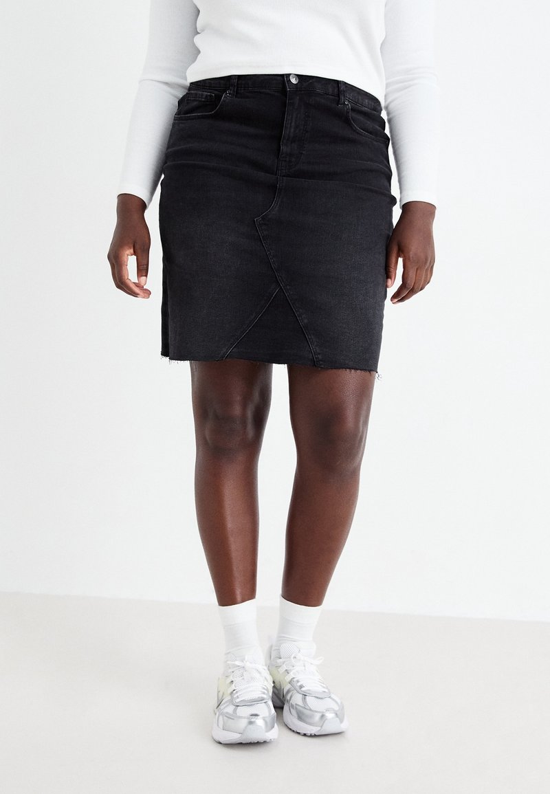 Černá denimová sukně s mírně třepeným lemem, předním zapínáním na patentky a diagonálními švy, doplněná šedými sportovními botami a bílými ponožkami.