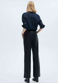 Pantaloni ampi in pelle nera con una texture liscia e due tasche posteriori, abbinati a una camicia di denim scuro con maniche arrotolate.