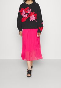 Svart stickad tröja med röd blommig broderi, i kombination med en genomskinlig rosa plisserad kjol och svarta slidessandaler.