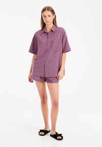 Camicia a maniche corte, con bottoni, tasca e motivo a scacchi piccoli in viola, abbinata a pantaloni corti coordinati e ciabatte nere.