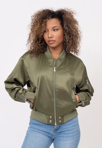 Olijfgroene satijnen bomberjack met ribgebreide mouwen en boord, borduursel op de borst en ritszakken op de mouwen. Aansluitend ontwerp.
