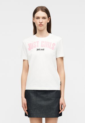 Γυναίκα που φοράει άσπρο t-shirt με ροζ γράμματα "JUST GIRLS" και μαύρα "Just Cavalli", συνδυασμένο με σκούρη μίνι φούστα τζιν.