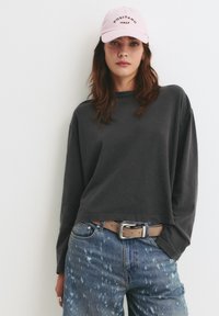 Grijze longsleeve shirt met een cropped ontwerp, gecombineerd met versleten blauwe denim jeans. Een roze pet met de tekst "POSITANO ITALY" maakt de outfit compleet.