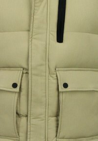 Beige gewatteerde bodywarmer met verticale ribbelstructuur, voorzien van een borstzak met rits en twee voorzakken met drukknopen in donker metaal.