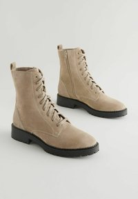 Botas de combate de ante beige con cordones en el frente y cremallera lateral, que presentan una suela texturizada y detalles de costura visibles.