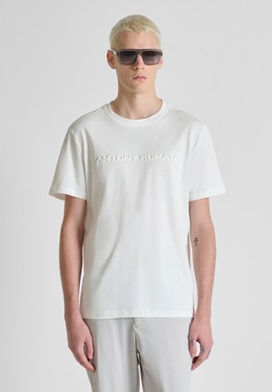 Giovane uomo con capelli biondi corti che indossa occhiali da sole neri oversize, maglietta bianca Antony Morato e pantaloni grigio chiaro, su sfondo grigio.