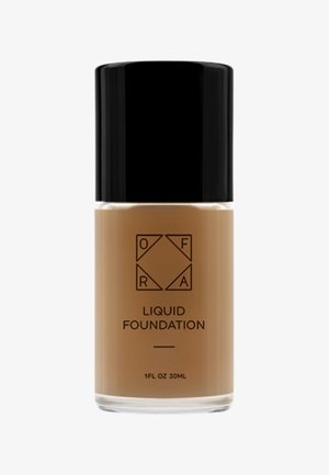 Braune Flüssigfoundation in einer klaren Glasflasche mit einem schwarzen Schraubverschluss. Auf der Vorderseite sind das Markenlogo und der Produkttext zu sehen.