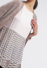 Lätt, stickad kofta i mjuk beige-grå, bärs över en mönstrad topp. Toppens övre del är i strukturerat vitt och den nedre delen är mönstrad i blått.