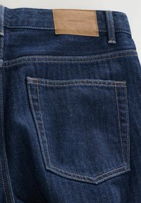 Granatowe dżinsy o gładkiej strukturze, z brązową skórzaną metką na pasie oraz jedną tylną kieszenią z kontrastującym przeszyciem.