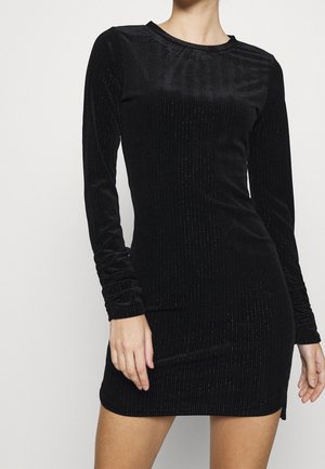 Femme portant une mini-robe noire ajustée à manches longues côtelées avec un léger brillant, debout devant un fond clair uni.