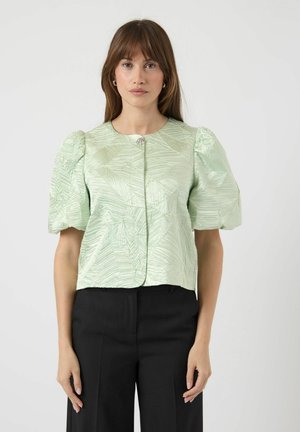 Vaaleanvihreä paita, jossa on teksturoitu lehtikuvio, lyhyet pussihihat, edessä napitus ja koristeellinen solki kaula-aukossa. Yhdistetty mustiin housuihin.