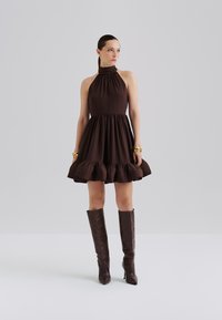 Brown halter-neck dress with a gathered waist and ruffled hem. Model wears sleek knee-high brown boots. Smooth fabric with a flowing silhouette.