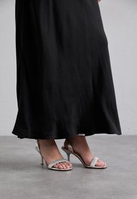 Sandale argintii cu toc înalt și două curele. Pantoful are o finisare lucioasă și un toc subțire stiletto, fiind asortat cu o rochie neagră fluidă.