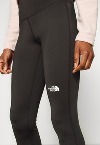 Mustat legginsit sileällä pinnalla, joissa on valkoinen logo reidessä ja kontrastiväriset saumat. Osa The North Face -brändistä.