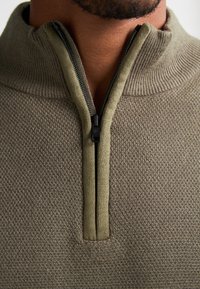 Pulover crem cu fermoar, cu un model de tricot texturat, având un guler înalt și un material contrastant în jurul fermoarului. Design casual și funcțional.