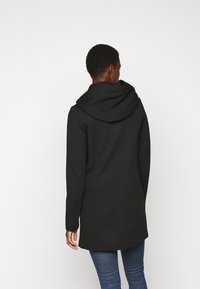 Must kollane kapuutsiga mantel, millel on kohandatud lõige, pikad varrukad ja sile tekstuur, kantud blue jeans'idega, millel on lihtne ja puhas disain.