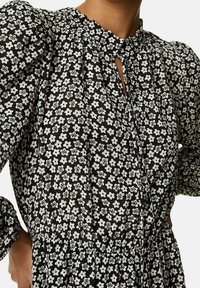 Vestido negro con estampado floral blanco, que presenta un escote alto, mangas abullonadas y una textura gather en la cintura. Detalle de botones en la parte delantera.