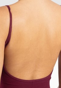 Vista trasera de una persona que lleva una camiseta sin mangas, acanalada, de color burdeos, con espalda descubierta, sobre un fondo blanco liso.