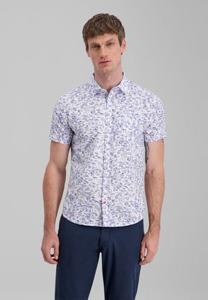 Man wearing a short-sleeve white button-up shirt with blue abstract patterns and dark blue pants standing against a plain gray background.