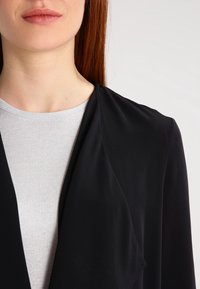 Blazer noir en tissu doux et fluide avec un col châle, superposé sur un top gris clair à côtes. Texture lisse, détails minimaux.