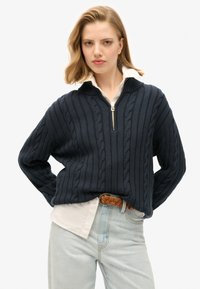 Granatowy sweter w warkocze z kołnierzem na zamek, teksturowanym wzorem i luźnym krojem. Nosi się go na białą koszulę z kołnierzykiem, w zestawieniu z jasnymi jeansami.