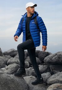Modrá bombera s kapucňou, čierna dlhá blúzka, čierne nohavice a čierne turistické topánky. Model stojí na skalnatom teréne. Viditeľná biela čiapka.