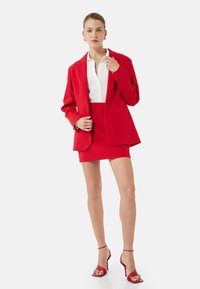 Rød skræddersyet blazer over en hvid skjorte, parret med en rød mini nederdel og røde højhælede sandaler. Glat stof, struktureret design og slank pasform.