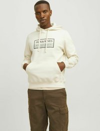 Krémová mikina s kapucí, přední kapsou, stahovací šňůrkou a černým logem na hrudi. V kombinaci s olivovými cargo kalhotami.