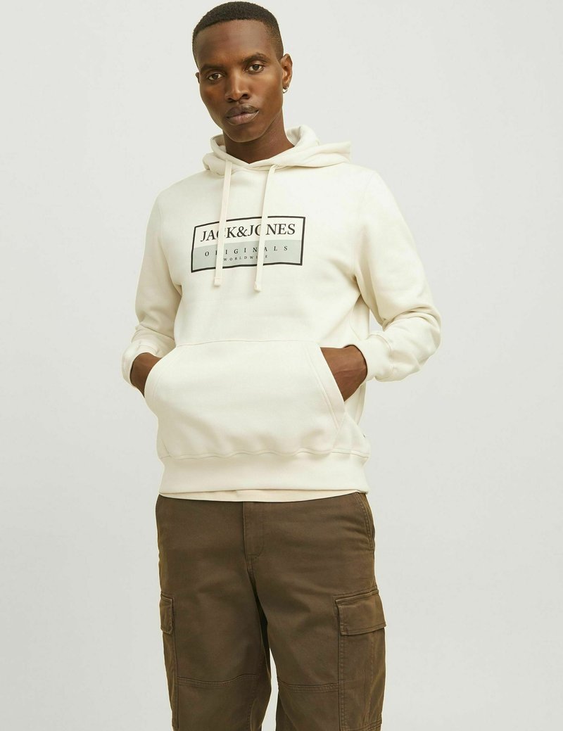 Krémová mikina s kapucí, přední kapsou, stahovací šňůrkou a černým logem na hrudi. V kombinaci s olivovými cargo kalhotami.