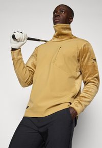 Sudadera de forro polar beige con cuello alto y mangas texturizadas, que presenta un bolsillo lateral y un acento de logotipo. Se lleva con pantalones oscuros y un guante de golf.
