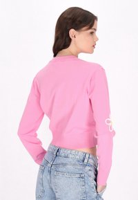 Maglione corto rosa con orlo a coste e maniche lunghe, che presenta un design floreale bianco e giallo sul braccio sinistro. Indossato con jeans a vita alta.