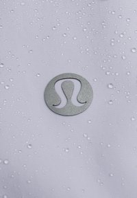 Vesikeelne valge kangas, millel on veepiisad, keskel metallikhall ringikujuline logo, millel on stiliseeritud "Lulu" disain.