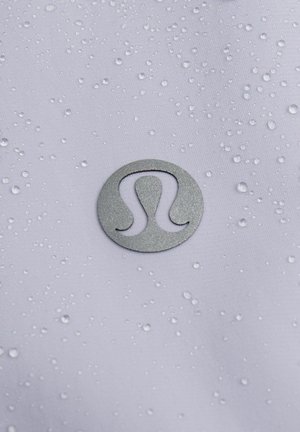 Waterdichte witte stof met druppels, met een metallic grijze cirkelvormige logo in het midden, dat een gestileerd "Lulu" ontwerp toont.