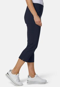 Marineblaue, knöchellange Hose mit taillierter Passform und seitlichen Schlitzen am Saum. Kombiniert mit weißen Sneakers mit silbernen Akzenten.