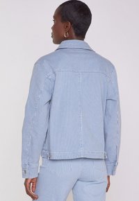 Giacca in denim a righe blu e bianche con colletto, chiusura con bottoni e vestibilità morbida, dotata di tasche laterali e dettagli sui polsini.
