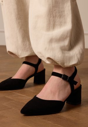 Scarpe nere in suede con tacco a blocco, punta appuntita e cinturino alla caviglia. L'outfit include pantaloni larghi color crema.