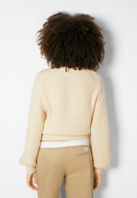 Vaalean beige ribattu neulepaita, jossa on lyhyt malli ja puffihihat, käytettynä beigejen housujen kanssa, nähtynä takaa.