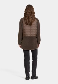 Braune Fleecejacke mit gestepptem Brustbereich. Verfügt über lange Ärmel, abgerundeten Saum und strukturierten Stoff. Getragen über schwarzen Jeans und Stiefeln.