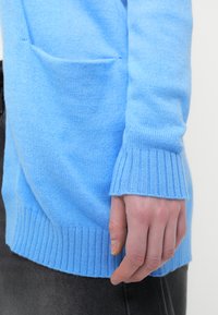 Ανοιχτό μπλε πλεκτό πουλόβερ με ριμπ μανσέτες και τελείωμα; διαθέτει μια τσέπη στο μπροστινό μέρος και λεία υφή. Εκτίθεται σε γκρι φόντο.