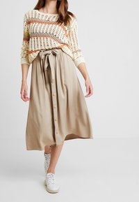 Pull léger en maille avec des rayures multicolores, associé à une jupe midi beige avec une ceinture à nouer et des boutons à l'avant. Porté avec des baskets blanches.