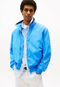 Blauwe nylon bomberjack met elastische manchetten, met het logo "TOMMY JEANS" op de borst. Gelaagd over een lichtgestreept overhemd.