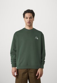 Sweatshirt vert en coton, avec un col rond et des poignets côtelés, arborant un petit motif graphique blanc sur la poitrine gauche.