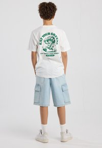 White cotton t-shirt with green graphic on back featuring a cartoon broccoli character and text; paired with light denim cargo shorts.