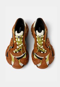 Zapatillas de malla marrones y blancas con suela de goma texturizada, acentos amarillos y un diseño ondulado único con bordes redondeados.