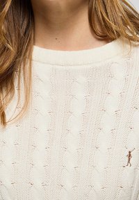 Gros plan sur une personne portant un pull crème à torsades avec une petite figure brodée en train de jouer au polo sur la poitrine.