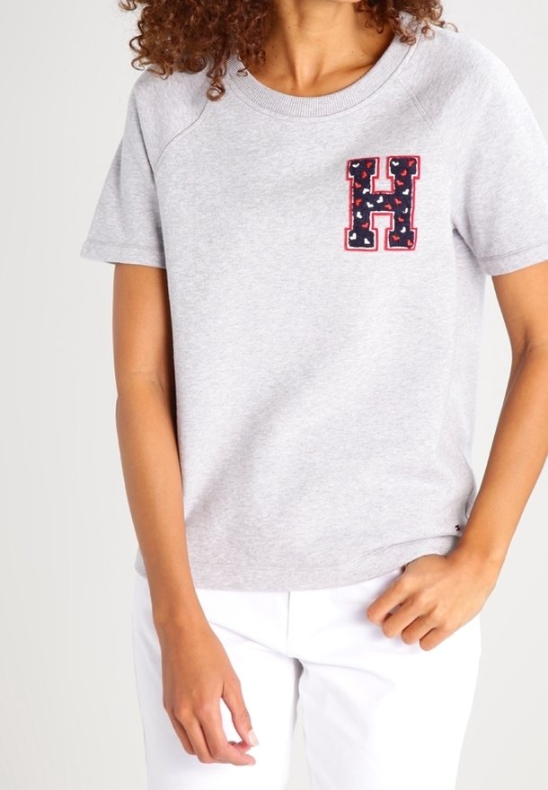 Ljusgrå kortärmad sweatshirt med rund halsringning, prydd med en dekorativ marinblå "H"-lapp med röd kant på bröstet.